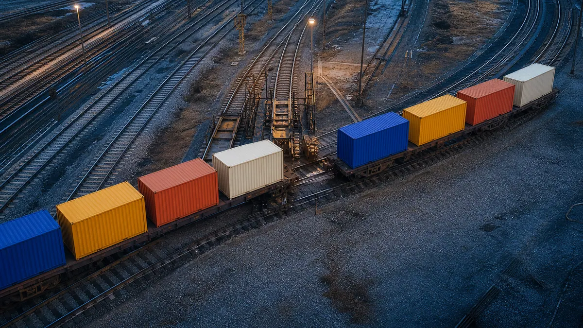 Unmarked blue, yellow, coral, and ivory freight containers cross a rail transfer junction in the same order at blue hour