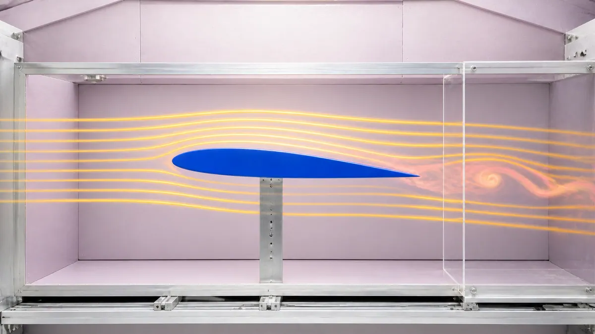 A cobalt airfoil in a pale-mauve wind tunnel, with saffron flow lines and a coral eddy behind a clear baffle.