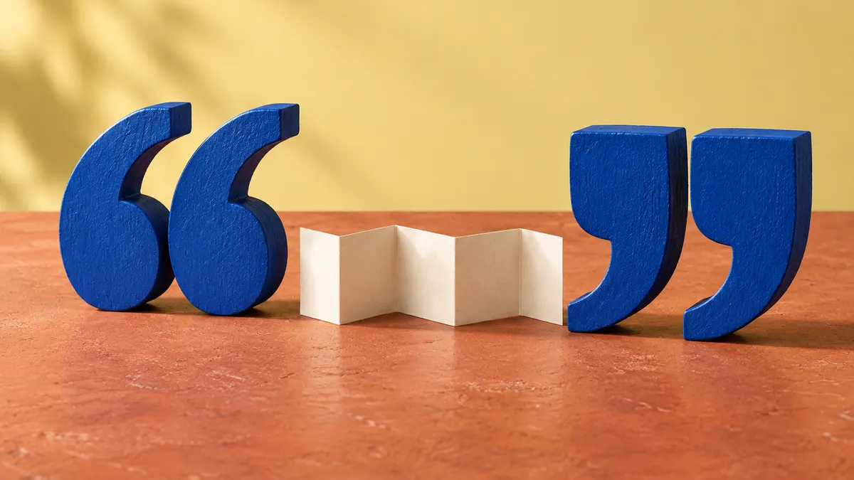 Cobalt wooden quotation marks frame a folded ivory paper strip on a terracotta tabletop.