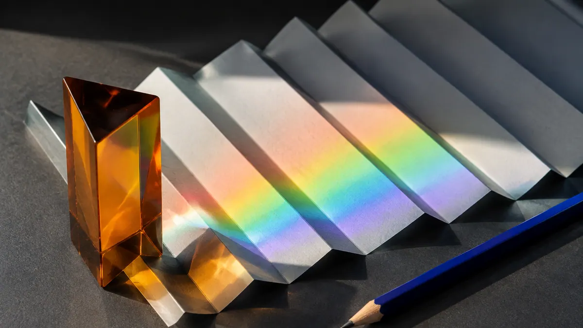 An amber prism casts a graduated spectrum across pleated white paper beside a cobalt pencil.