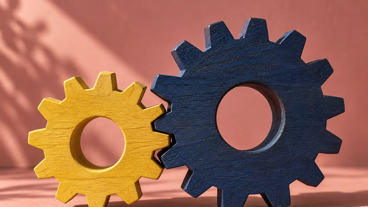 A yellow wooden gear meshes with a larger indigo wooden gear on a warm pink surface.