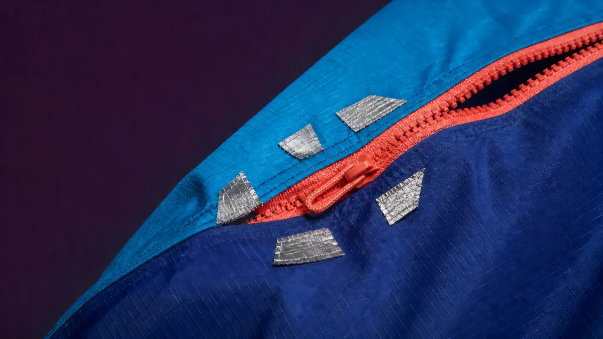 A coral zipper with a braided cord pull closes a jagged opening in layered blue technical fabric under raking studio light.